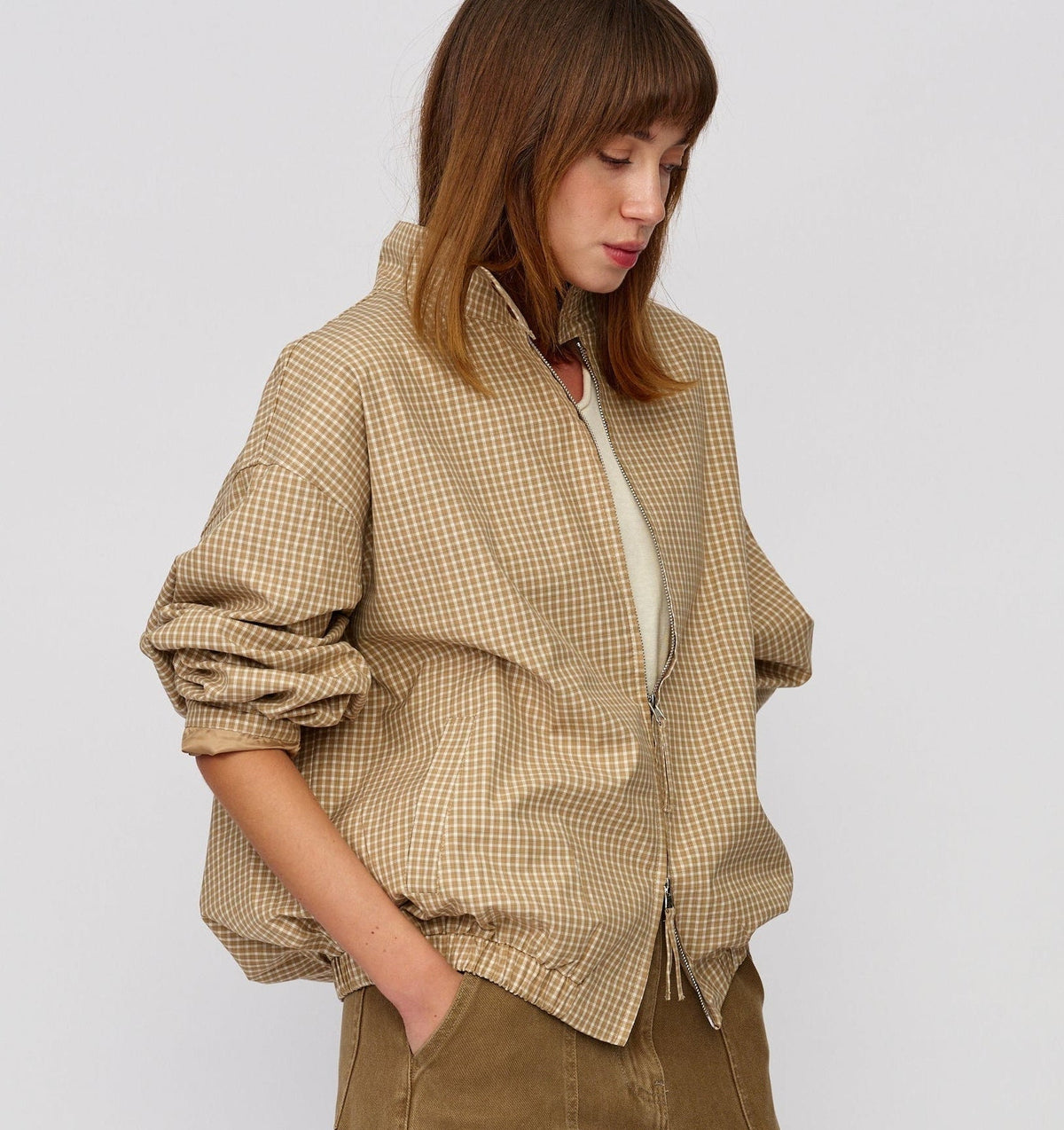 A woman wearing a beige and brown gingham overshirt with rolled-up sleeves, layered over a cream top and paired with brown trousers, photographed against a plain light gray studio background.